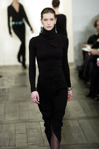 A model walks a fashion runway wearing a fitted black turtleneck top and black pleated skirt with front buttons.
