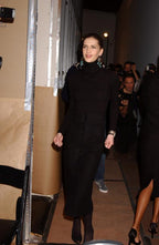 A woman walking in a backstage corridor wearing a long black turtleneck dress with front buttons and black pointed shoes. Photographers and other people are visible in the background.