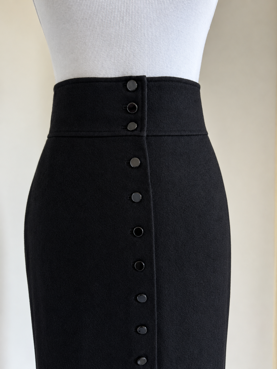 A close-up of a black high-waisted pencil skirt on a white mannequin torso, featuring a prominent vertical row of dark buttons down the front center.