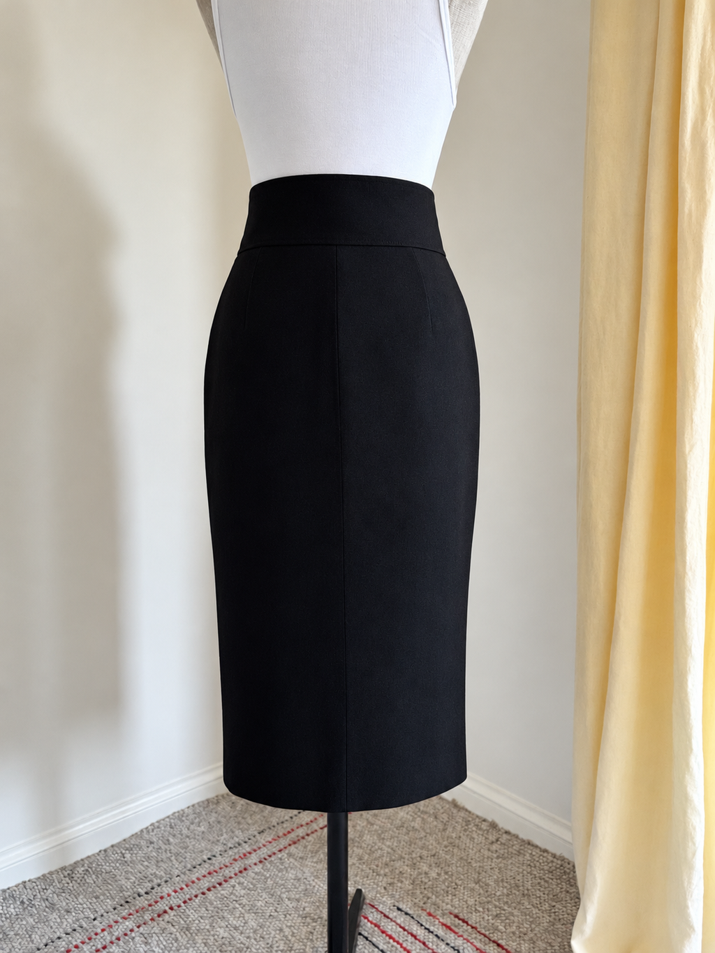 A black high-waisted pencil skirt displayed on a white dress form mannequin, set against an indoor background with a cream wall, a yellow curtain, and a patterned carpet.
