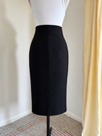 A black high-waisted pencil skirt displayed on a white dress form mannequin, set against an indoor background with a cream wall, a yellow curtain, and a patterned carpet.