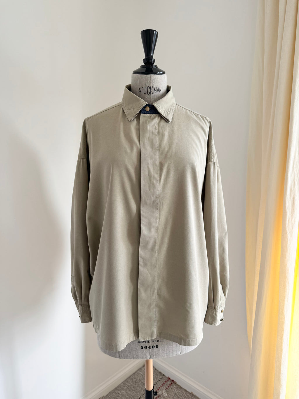 A light beige long-sleeve button-up shirt displayed on a dress form mannequin in an indoor setting with a white wall and a beige curtain on the right.