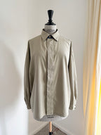 A light beige long-sleeve button-up shirt displayed on a dress form mannequin in an indoor setting with a white wall and a beige curtain on the right.