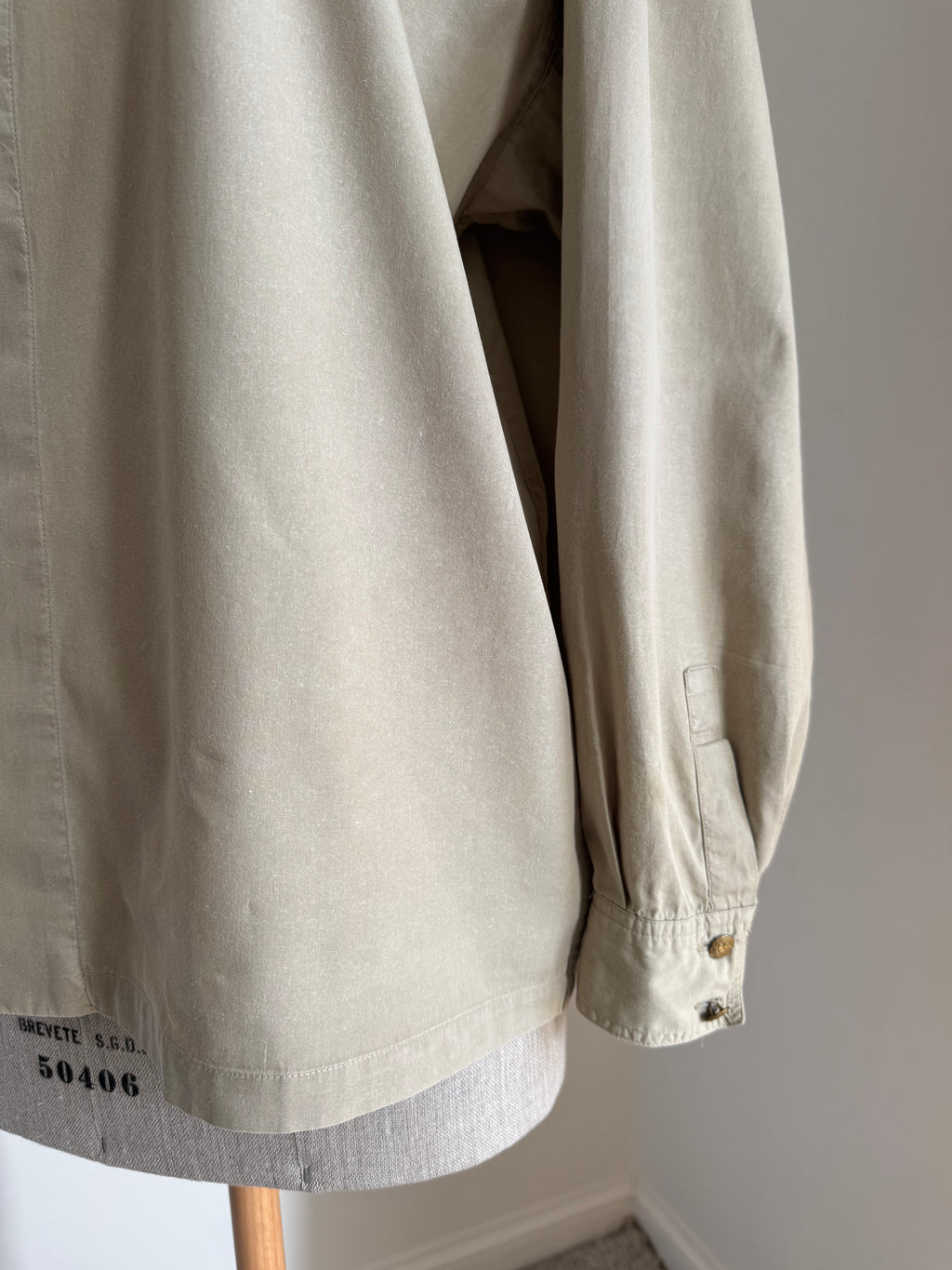 A close-up of a light beige cotton overshirt or jacket hanging on a wooden hanger against a plain light gray wall. The sleeve is shown with buttoned cuffs, and a small black printed code is visible near the lower left hem.