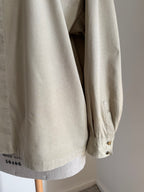 A close-up of a light beige cotton overshirt or jacket hanging on a wooden hanger against a plain light gray wall. The sleeve is shown with buttoned cuffs, and a small black printed code is visible near the lower left hem.