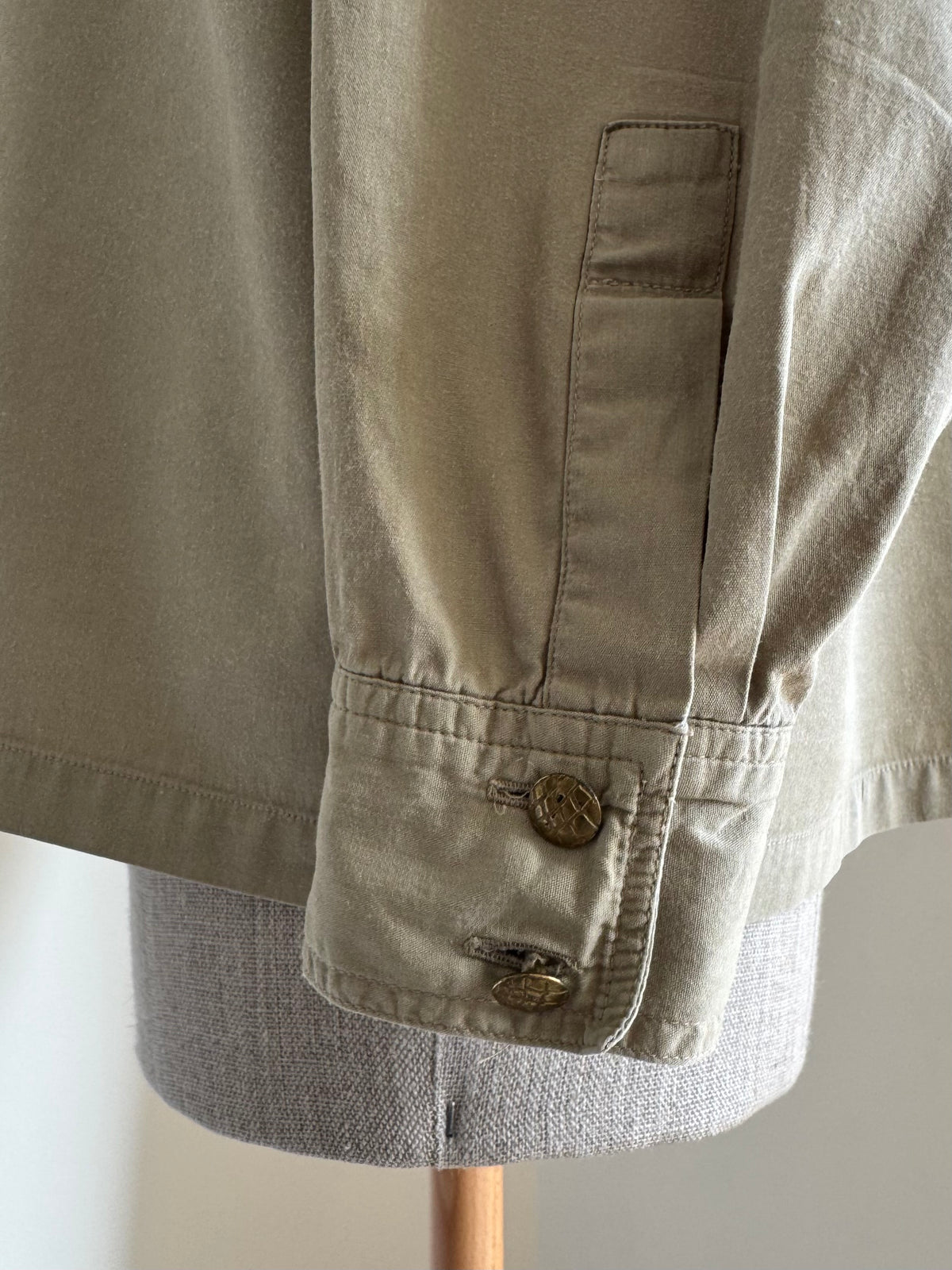 Close-up of a light beige or khaki jacket or overshirt displayed on a gray dress form, showing the buttoned cuff detail.