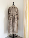 A plush, textured bathrobe in a taupe-beige color displayed on a black dress form mannequin in a home setting with a white wall and cream curtains.