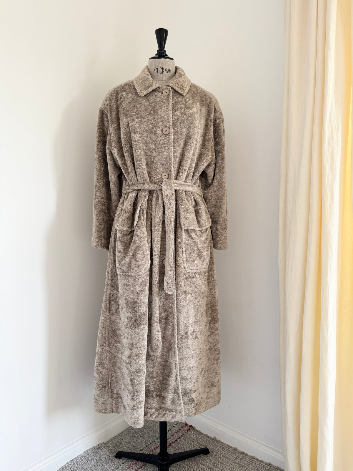 A plush, textured bathrobe in a taupe-beige color displayed on a black dress form mannequin in a home setting with a white wall and cream curtains.