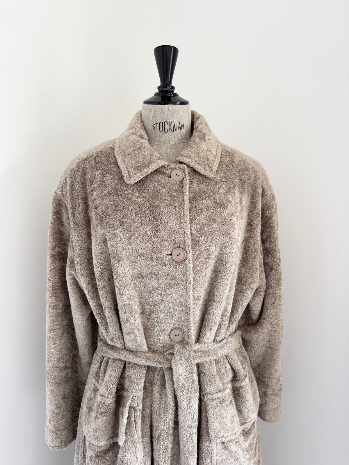 A plush, textured beige bathrobe with a collar, button closure, and matching waist tie, displayed on a mannequin torso against a plain white wall.