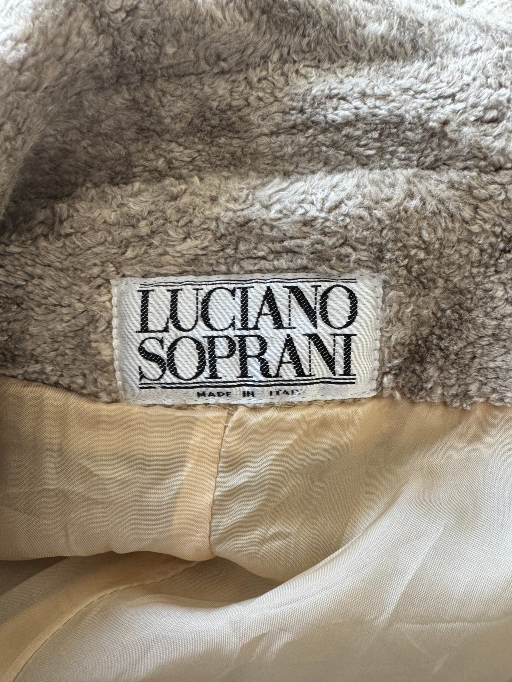 Close-up of a plush taupe-gray fleece lining with a sewn-on brand label reading 'LUCIANO SOPRANI' on a cream-colored garment.