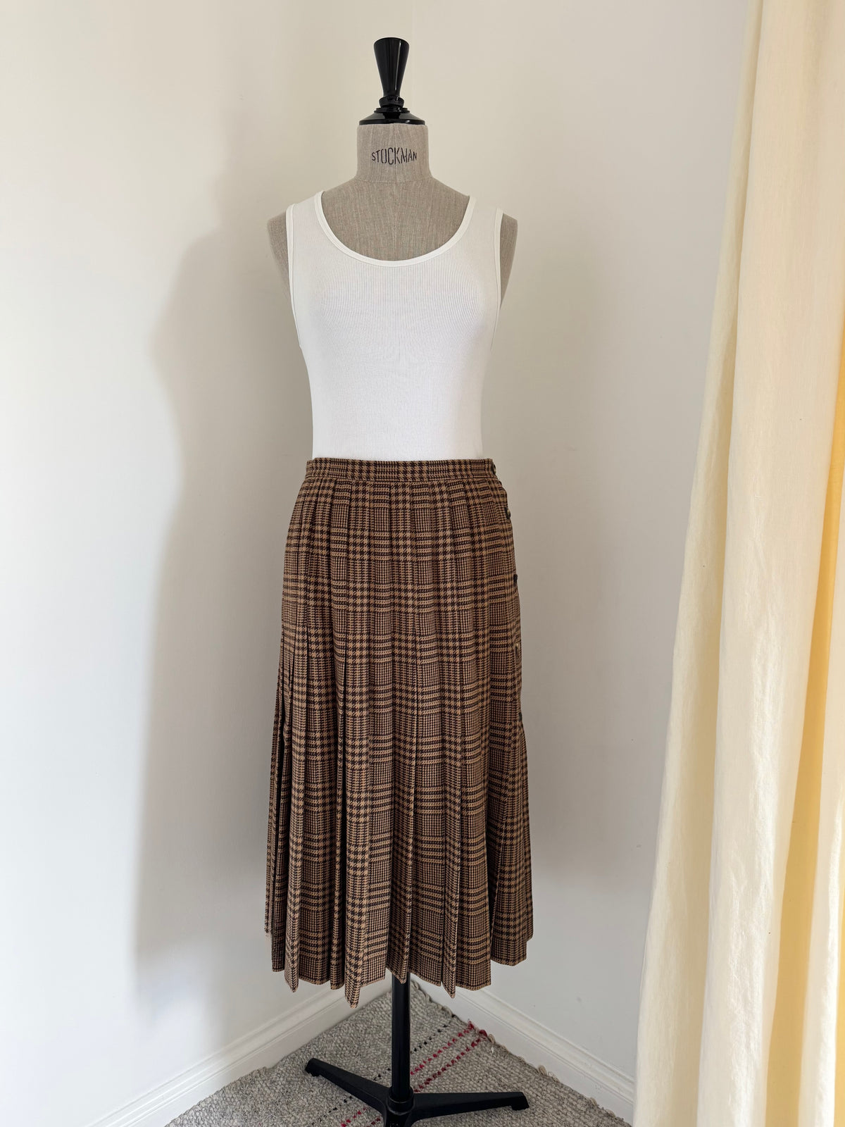 A brown and beige vintage valentino plaid pleated midi skirt displayed on a dress form mannequin, paired with a white ribbed tank top, in a home setting with a white wall and a cream curtain.