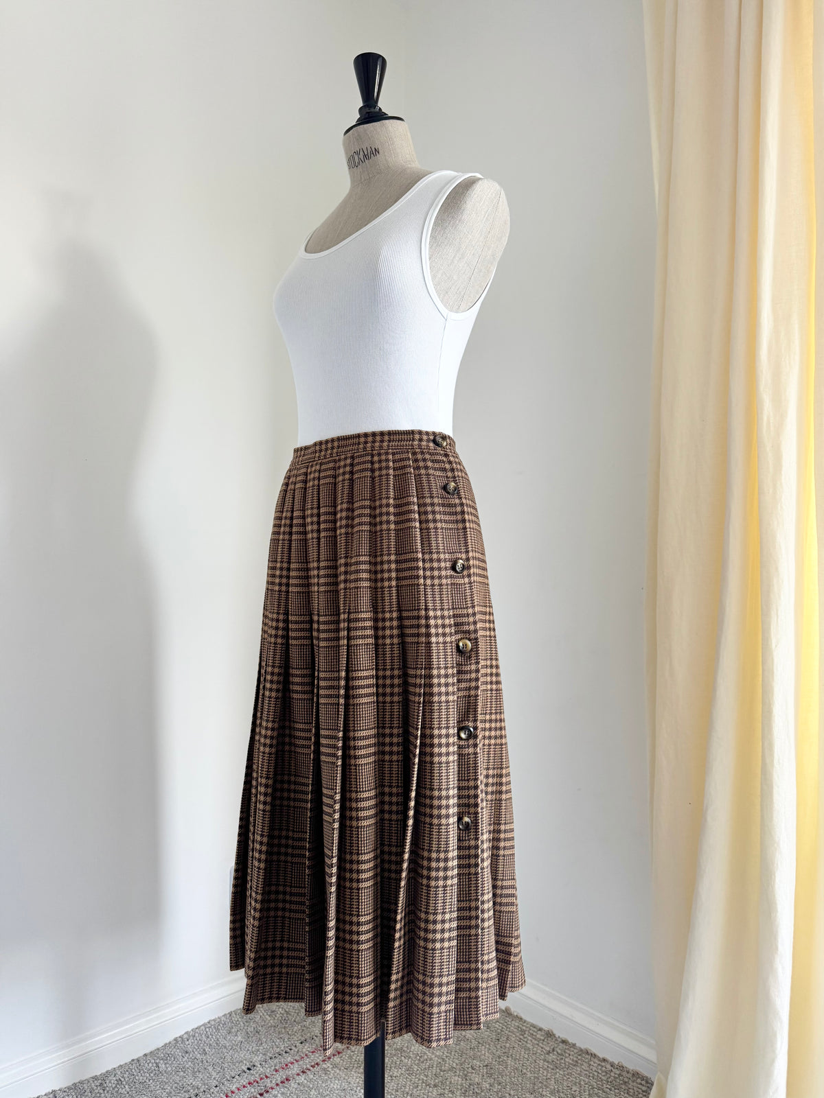 A brown and beige Valentino plaid pleated midi skirt with a button-down front, displayed on a mannequin wearing a white ribbed tank top in a room corner.