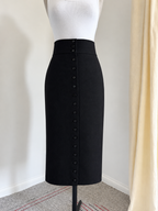 A black high-waisted pencil skirt with a vertical row of buttons down the front, displayed on a white mannequin torso in an indoor setting.
