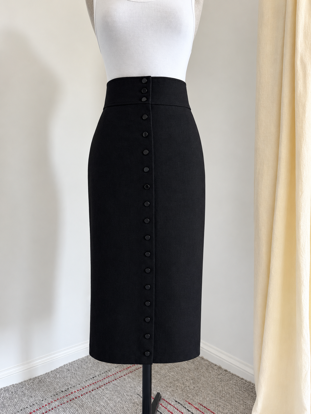 A black high-waisted pencil skirt with a vertical row of buttons down the front, displayed on a white mannequin torso in an indoor setting.